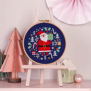 Santa themed cross stitch piece framed in embroidery hoop on shelf with other Christmas decorations