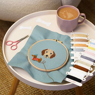 Dog themed cross stitch design in embroidery hoop placed on table next to threads, scissors and cup of tea