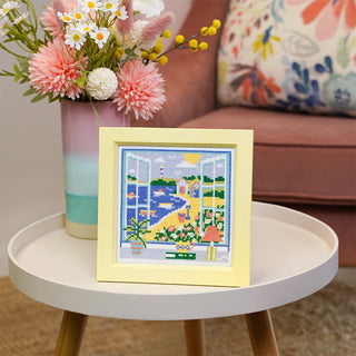 Summer View stitched sample in frame on table with vase of flowers